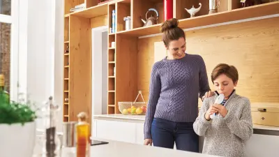 Mother with son standing in kitchen, son blowing in Vivatmo me.