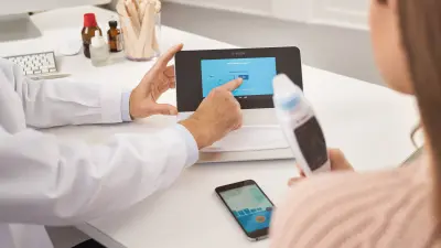 Patient holding Vivatmo meter in her hand. Vivatmo pro lying on doctor's desk in front of her. Doctor points to the screen with the Finder.